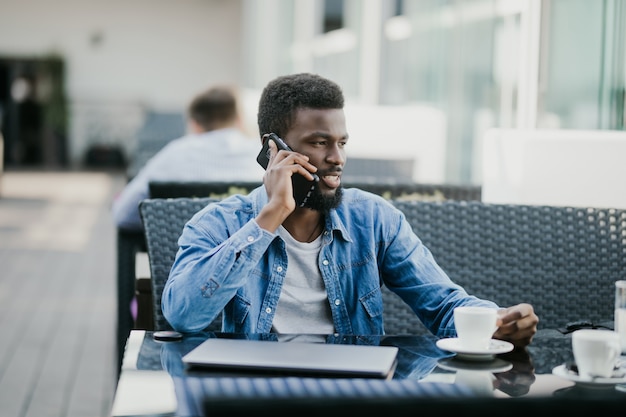 Zdjęcie uśmiechnięty młody człowiek afryki picia kawy i rozmawia przez telefon komórkowy, siedząc w kawiarni