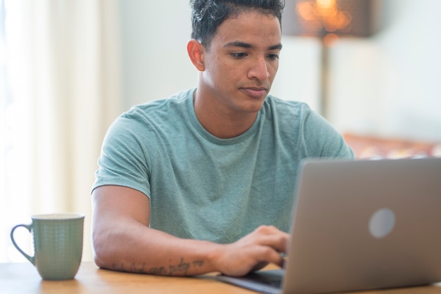 Uśmiechnięty młody człowiek freelancer za pomocą laptopa studiujący online, pracujący w domu, szczęśliwy przypadkowy hipster facet piszący na komputerze notebook surfowanie po Internecie patrząc na ekran korzystający z odległej pracy siedzieć przy stole