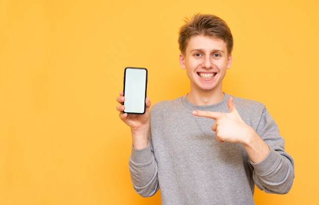 Uśmiechnięty młody człowiek stoi na kolorze żółtym i pokazuje palec na smartphone