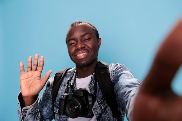 Uśmiechnięty młody entuzjasta fotografii posiadający lustrzankę cyfrową i biorąc selfie podczas machania do aparatu na niebieskim tle. Pewny siebie fotograf z profesjonalnym aparatem fotograficznym i plecakiem.