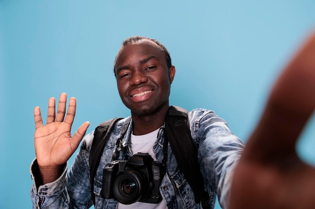 Uśmiechnięty Młody Entuzjasta Fotografii Posiadający Lustrzankę Cyfrową I Biorąc Selfie Podczas Machania Do Aparatu Na Niebieskim Tle. Pewny Siebie Fotograf Z Profesjonalnym Aparatem Fotograficznym I Plecakiem.