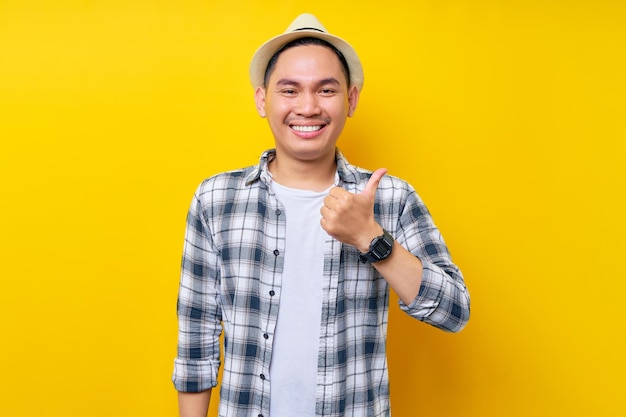 Uśmiechnięty młody przystojny etniczny Azjata, 20-latek ubrany w przypadkowe ubrania, kapelusz, stojący pewnie i pokazujący kciuk w górę jak gest odizolowany na żółtym tle Koncepcja stylu życia ludzi