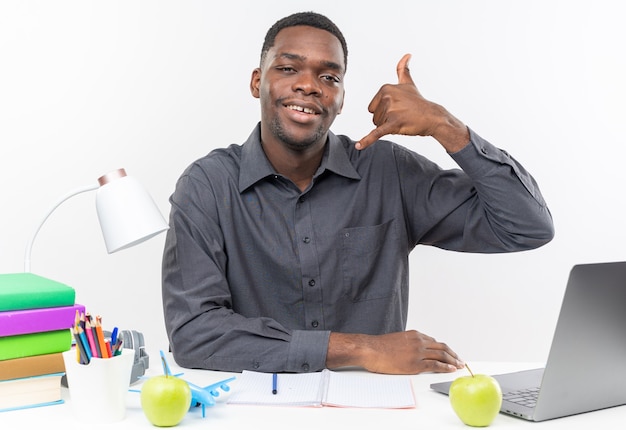 Uśmiechnięty młody student afroamerykański siedzący przy biurku z szkolnymi narzędziami gestykuluje zadzwoń do mnie znak