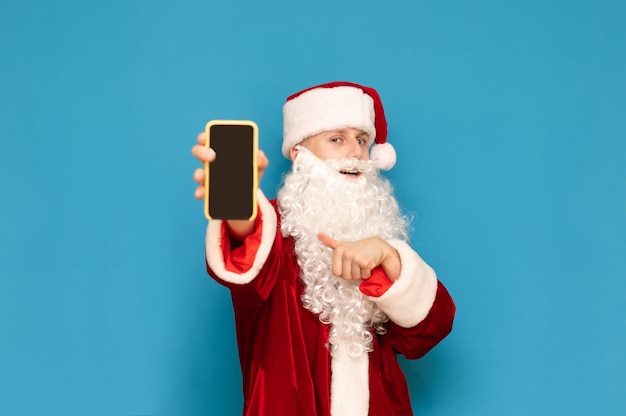 Uśmiechnięty Młody święty Mikołaj Z Smartphone W Ręce Na Błękicie, Pokazuje Telefon Z Czerń Ekranem W Kamerze I Pokazuje Palec