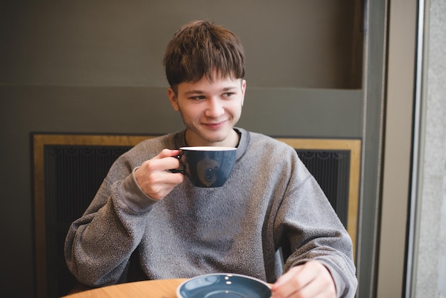Uśmiechnięty nastolatek z pokolenia Z 1617 lat pijący świeżą smaczną kawę siedzący w kawiarni w pomieszczeniu Przystojny blond nastoletni hipster