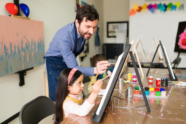 Uśmiechnięty nauczyciel chętnie pomaga małej dziewczynce malować na płótnie podczas zajęć plastycznych dla dzieci