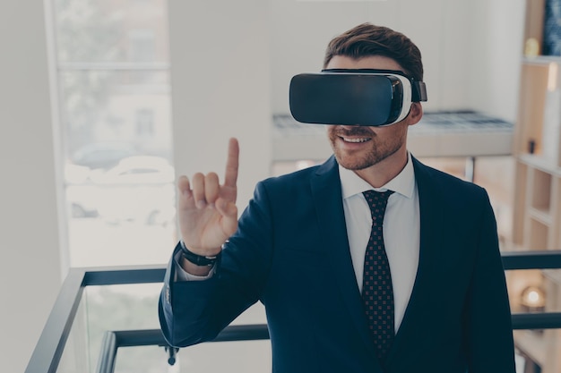 Uśmiechnięty odnoszący sukcesy biznesmen w garniturze stojący we wnętrzu biura i używający okularów VR do pracy