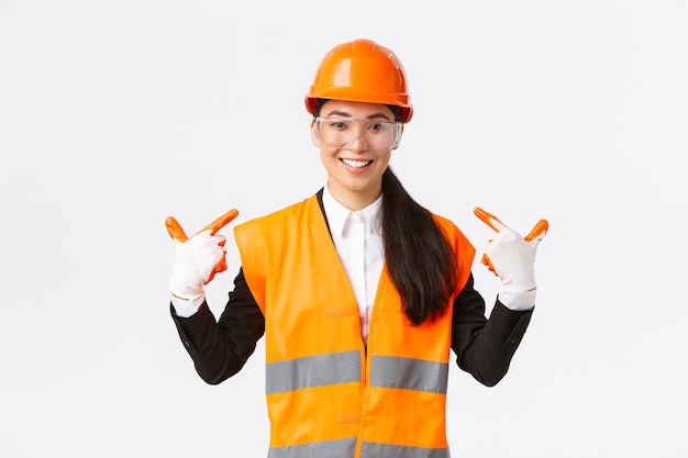 Uśmiechnięty pewny siebie azjatycki główny inżynier budowlany w kasku ochronnym, rękawiczkach i okularach wskazujących na siebie, powiadomić o przestrzeganiu bezpiecznego protokołu i nosić specjalną odzież, białe tło