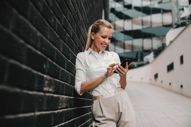 Uśmiechnięty piękny modny bizneswoman stojący na zewnątrz i za pomocą inteligentnego telefonu. Na zewnątrz centrum biznesowego.