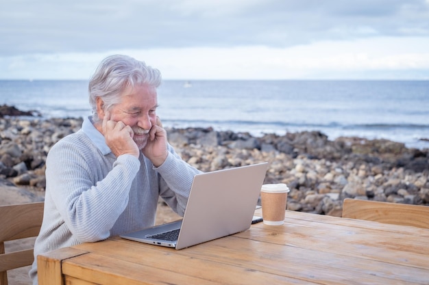 Uśmiechnięty piękny starszy mężczyzna siedzący na świeżym powietrzu przy drewnianym stole za pomocą laptopa w alternatywnym biurze
