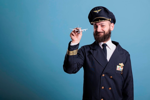 Uśmiechnięty pilot w mundurze trzymający widok z przodu małego modelu samolotu, lotnik z akademii lotniczej bawiący się zabawką samolotu komercyjnego. Członek załogi samolotu patrzący w kamerę, średnie ujęcie studyjne