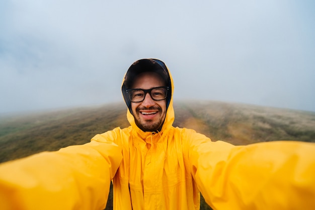 Uśmiechnięty podróżnika mężczyzna w żółtym płaszczu bierze selfie w mgłowym dniu na polu