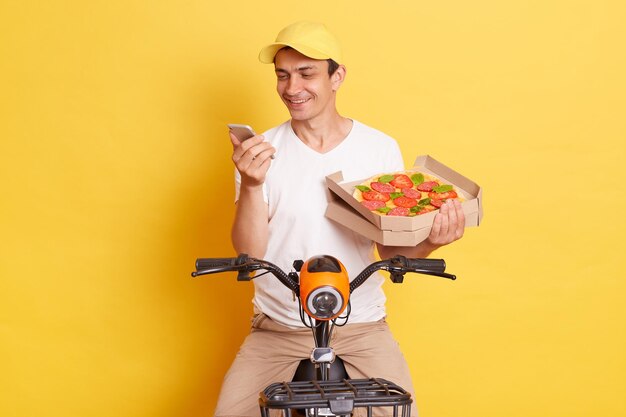 Uśmiechnięty pozytywny dostawca ze świeżo upieczoną pizzą w kartonowym pudełku, ubrany w białą koszulkę, siedzący na szybkim motocyklu i używający telefonu komórkowego, czekając na klienta odizolowanego na żółtym tle