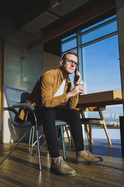Uśmiechnięty pracownik płci męskiej w przypadkowych ubraniach odwraca wzrok podczas rozmowy przez telefon komórkowy i picia kawy podczas przerwy w nowoczesnym kreatywnym miejscu pracy
