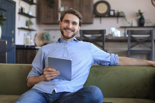 Uśmiechnięty przystojny mężczyzna trzymający i używający tabletu z sieciami społecznościowymi w domu, szczęśliwy freelancer robiący zakupy online, siedzący na wygodnej kanapie w salonie