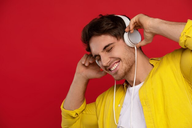 uśmiechnięty przystojny mężczyzna w żółtej koszuli słuchania muzyki w słuchawkach z zamkniętymi oczami, odizolowane na czerwono