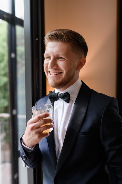 Uśmiechnięty przystojny pan młody w stylowym garniturze z krawatem motylkowym trzymającym szklankę z napojem stojącym w pobliżu dużego okna Portret eleganckiego narzeczonego czekającego na ceremonię ślubną