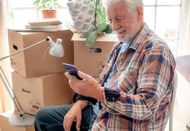 Uśmiechnięty przystojny starszy mężczyzna w dniu przeprowadzki siedzi odpoczywając patrząc na kartonowe pudełka na telefony komórkowe