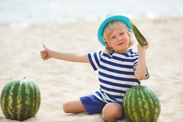 Uśmiechnięty Rozochocony Dziecko Z Arbuzem Na Dennym Brzeg. ładny Mały Chłopiec Na Plaży Jedzenie Arbuza. Szczęśliwy Uśmiechnięty Dziecko Kciuk W Górę