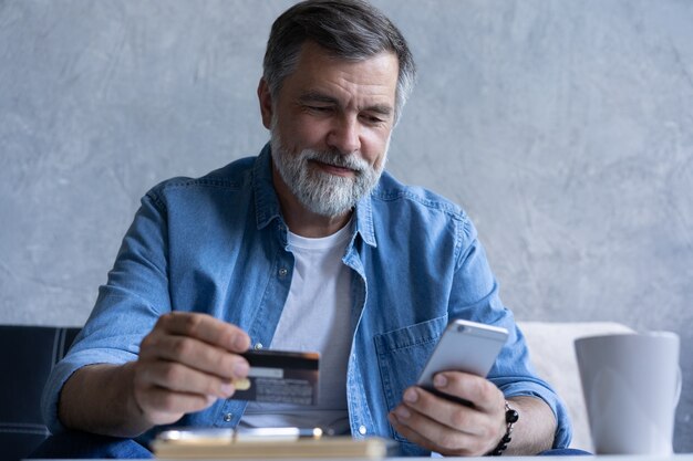 Uśmiechnięty siwy mężczyzna z lat 50-tych płacić rachunki online za pomocą gadżetu nowoczesnego smartfona i karty kredytowej. Szczęśliwy dojrzały klient banku dokonać zakupu płatności internetowych na telefon komórkowy z domu. Koncepcja technologii.