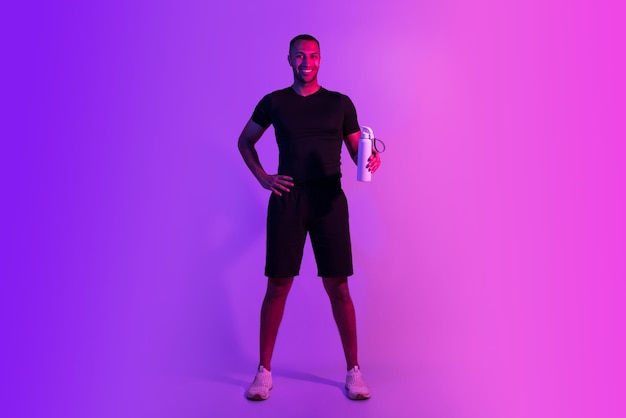 Uśmiechnięty sportowy czarny facet trzyma butelkę wody na fioletowym tle