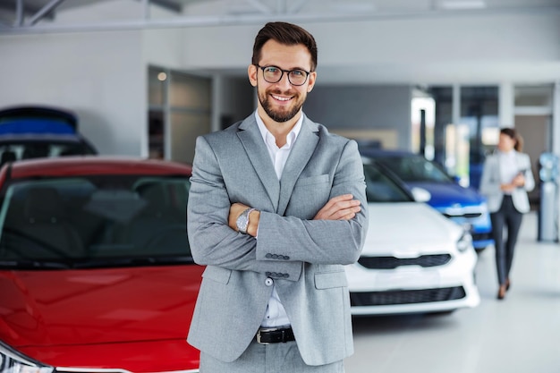 Uśmiechnięty sprzedawca samochodów stojący w salonie samochodowym z skrzyżowanymi rękami i czekający na klientów, którzy przyjdą do sprzedaży