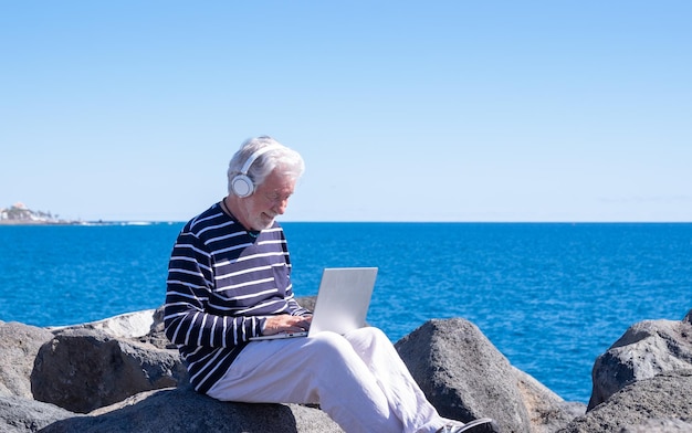 Uśmiechnięty starszy mężczyzna rasy kaukaskiej ze słuchawkami siedzący na zewnątrz na morzu, korzystający z laptopa w pracy zdalnej, siwowłosy starszy mężczyzna przeglądający z komputerem