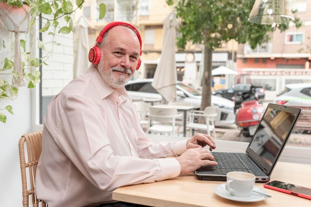 Uśmiechnięty starszy mężczyzna siedzący w kawiarni ze słuchawkami używa laptopa do nauki języków, lekcje online, ciesząc się emeryturą patrząc w kamerę