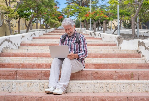 Uśmiechnięty starszy mężczyzna w kraciastej koszuli siedzący na zewnątrz na schodach w mieście za pomocą laptopa Brodaty mężczyzna rasy kaukaskiej uzależniony od technologii i społeczności