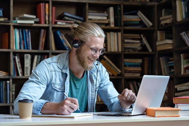 Uśmiechnięty student nosi zestaw słuchawkowy i patrzy na laptopa, robiąc notatki w bibliotece