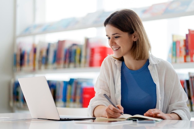 Uśmiechnięty student uniwersytetu studiujący, robiący notatki za pomocą laptopa w bibliotece. Koncepcja edukacji