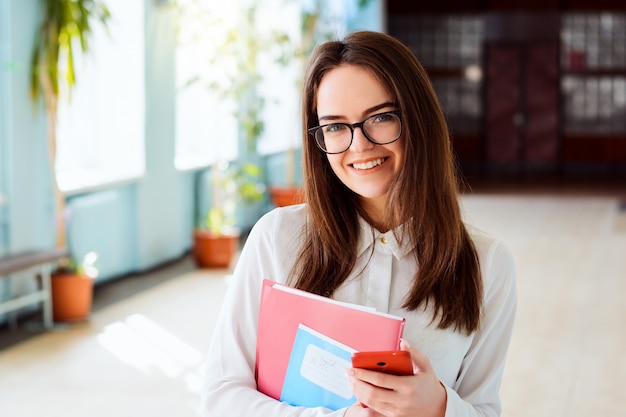 Uśmiechnięty student w okularach z książkami, zeszytami i telefonem komórkowym w budynku uniwersytetu