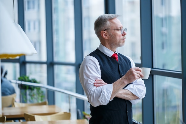 Uśmiechnięty szczęśliwy dyrektor zarządzający myśli o pomyślnym rozwoju kariery, stojąc z filiżanką kawy w dłoni w swoim biurze w pobliżu okna z miejscem na kopię