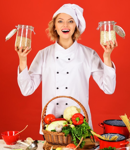 Zdjęcie uśmiechnięty szef kuchni w białym mundurze stoi przy stole z warzywami w koszyku i trzyma szklane słoiki z mąką