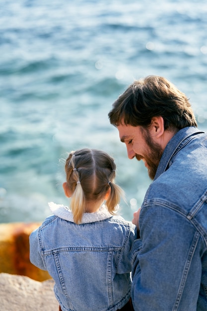 Uśmiechnięty tata siedzi obok małej dziewczynki na kamieniu nad zbliżeniem widoku z tyłu na morze
