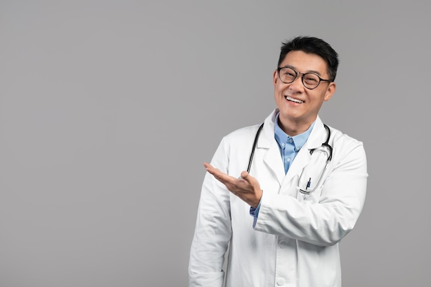 Uśmiechnięty terapeuta dorosłego koreańskiego mężczyzny w białych okularach ze stetoskopem pokazuje rękę na pustej przestrzeni