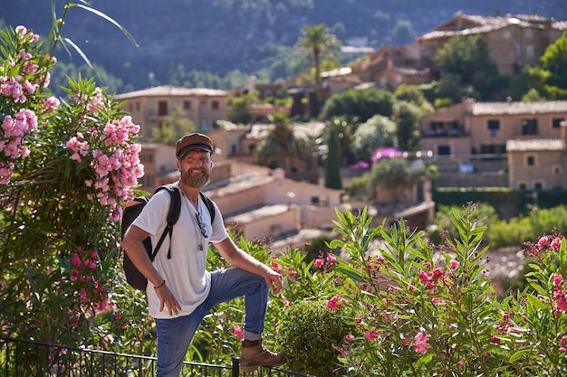 Uśmiechnięty turysta płci męskiej w stylowym stroju z plecakiem w pobliżu kwitnących krzaków i cieszący się widokiem na stare miasto w letni dzień