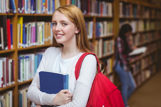 Uśmiechnięty uczeń z plecakiem trzyma książkę w bibliotece