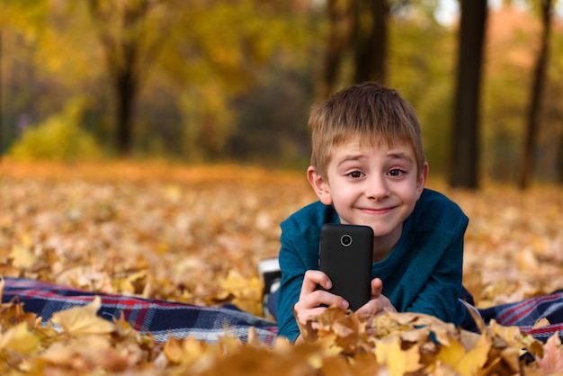 Uśmiechnięty uczeń ze smartfonem leżącym na żółtych jesiennych liściach w kratę jesienny dzień