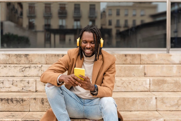 Uśmiechnięty współczesny mężczyzna w bezprzewodowych żółtych słuchawkach słuchający muzyki i siedzący na schodach podczas korzystania ze smartfona