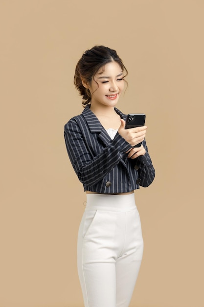 Uśmiechnij się piękna azjatycka biznesowa kobieta trzymająca telefon komórkowy na odosobnionym beżowym tle kopii przestrzeni
