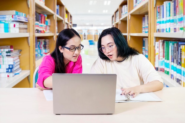 Uśmiechnijcie się do młodych ludzi patrzących na ekran laptopa, komunikujących się z dwiema studentkami przy biurku, korzystających z laptopa z półką na książki w tle biblioteki