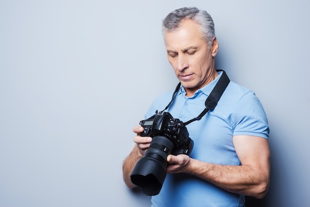 Ustawianie aparatu. Portret starszego dojrzałego mężczyzny w koszulce trzymającego aparat stojący na szarym tle