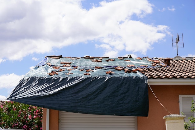 Uszkodzony przez burzę dach na domu z czarną plastikową plandeką nad dziurą w gontach i dachu po gwałtownej wiosenno-letniej burzy