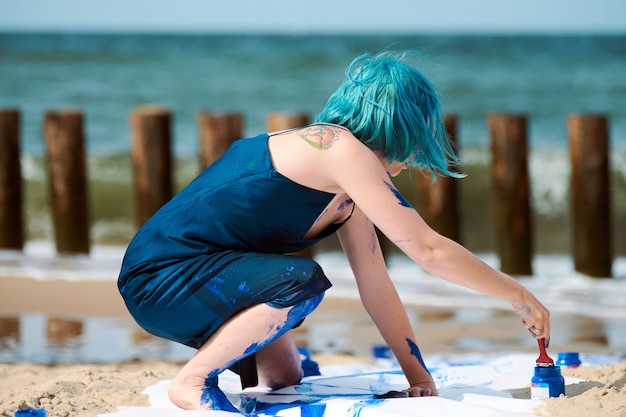 Utalentowana niebieskowłosa artystka performerka w ciemnoniebieskiej sukience posmarowanej niebieskim gwaszem z szerokimi pociągnięciami na dużym płótnie na plaży. Ekspresyjna sztuka performance, sztuka współczesna w plenerze