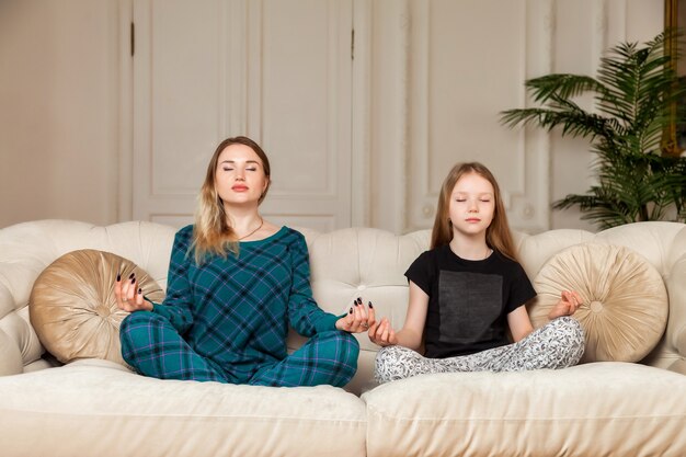 Uważna Szczęśliwa Mama Z Uroczą Zabawną Córką Robi ćwiczenia Jogi W Domu W Salonie, Mama I Mała Dziewczynka Razem Siedząc W Pozycji Lotosu Na Kanapie, Mama Uczy Dziecko Medytacji. Skopiuj Miejsce