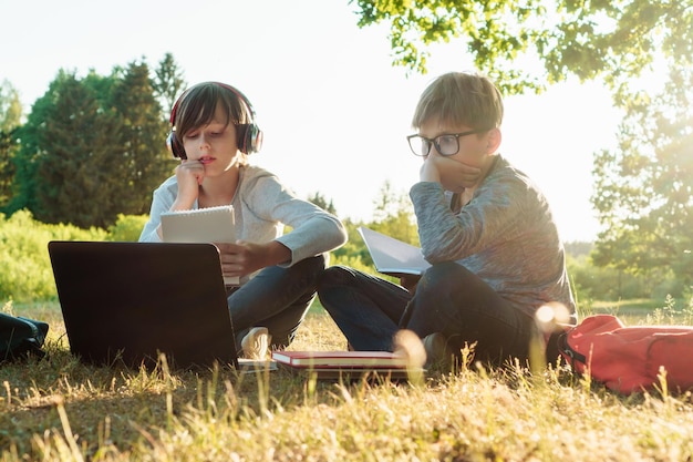 Uważny uczeń w okularach patrzący na monitor laptopa i opierając głowę na dłoni, siedząc obok kolegi z klasy w słuchawkach, który czyta lekcję z zeszytu ćwiczeń Dwóch kolegów ze szkoły studiujących w parku