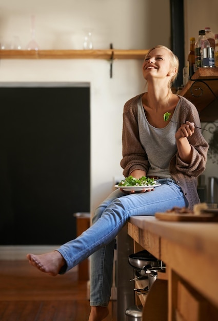 Uwielbia swoją nową dietę Ujęcie atrakcyjnej młodej kobiety siedzącej na kuchennym blacie, śmiejącej się podczas jedzenia sałatki