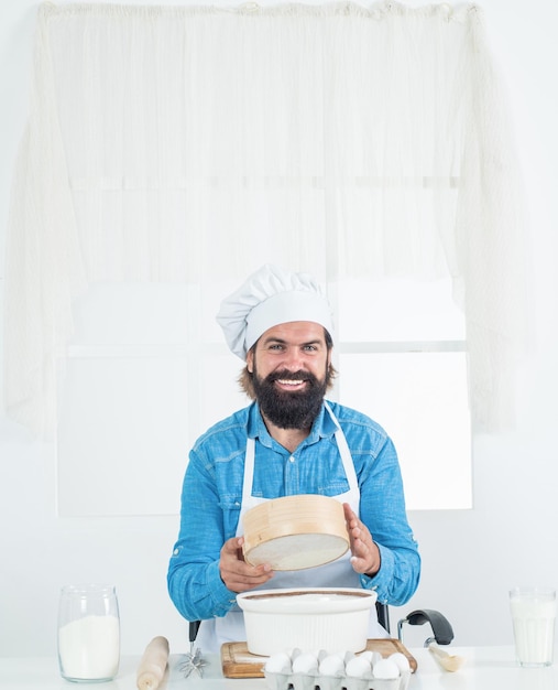 Uwielbiam moją pracę dojrzały brodaty mężczyzna nosi kapelusz szefa kuchni za pomocą sita mężczyzna kucharz przygotowuje jedzenie w domowej kuchni brutalny hipster w fartuchu gotowanie czas posiłku do jedzenia szczęśliwego pieczenia koncepcja sprzątania