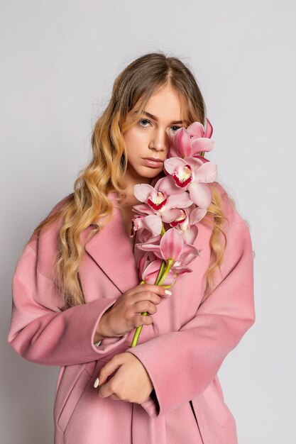 Uwodzicielska blond kobieta w różowej kurtce pozowanie Studio na szarym tle. Moda piękny portret elegancki model w pastelowym stroju dorywczo wiosna. Piękna dziewczyna o zdrowej skórze i gałęzi orchidei